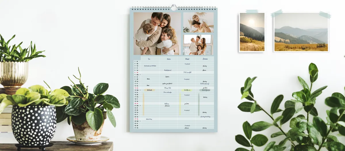 Familienkalender mit Fotos einer fröhlichen Familie aus Mutter, Vater und zwei Kindern hängt an der Wand, ringsherum stehen Pflanzen