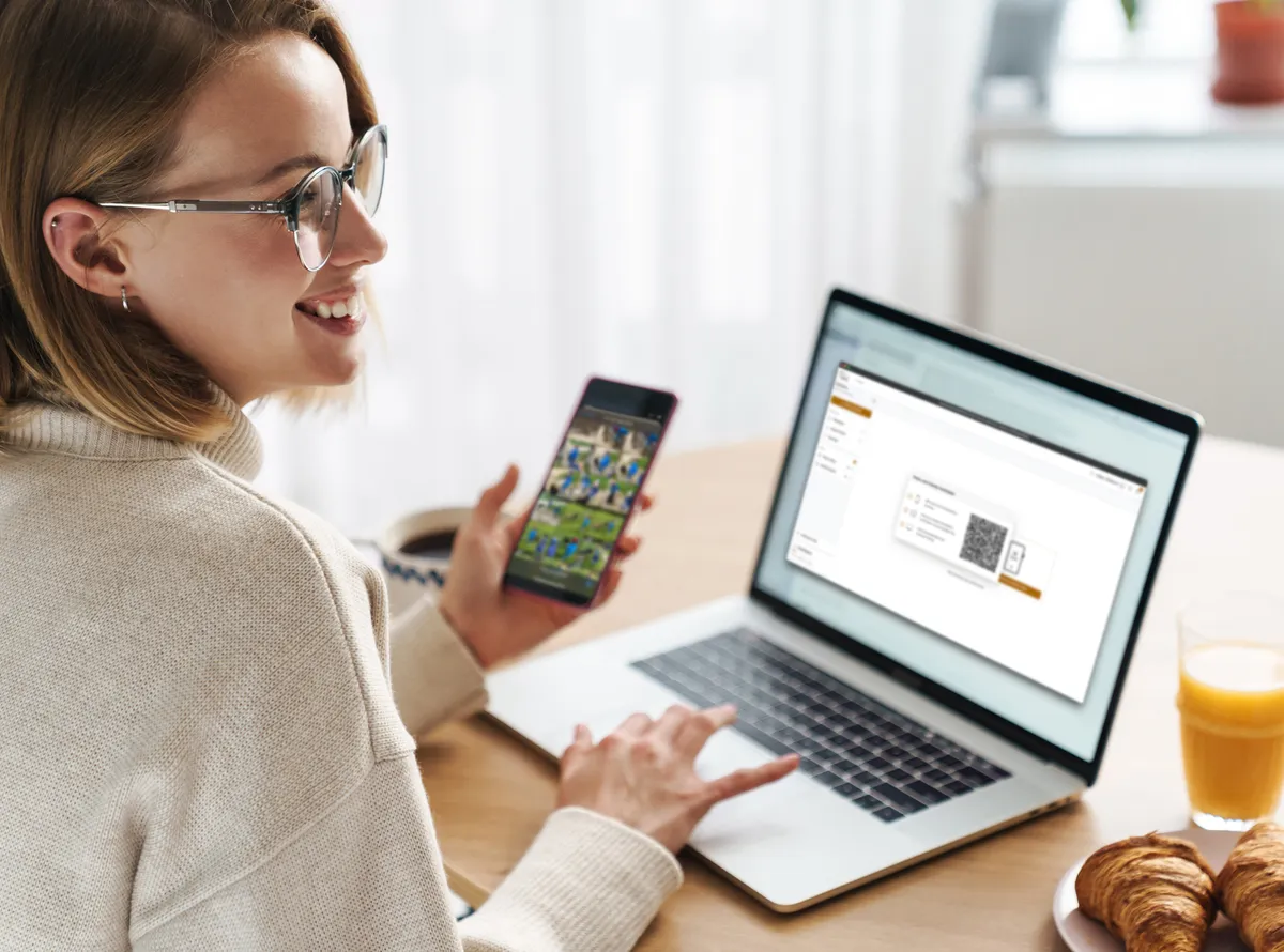 Lächelnde Frau am Laptop hält ihr Smartphone in der Hand und lädt über einen QR-Code easy ihre Bilder vom Smartphone auf das Laptop